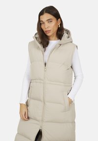 Gilet imbottito beige con cappuccio, chiusura con zip, tasche laterali e lunghezza lunga. Texture morbida e dettaglio del design trapuntato.