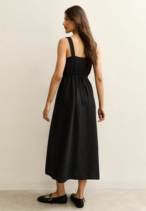 Zwarte maxi-jurk met een getextureerde stof, brede bandjes en een taille met trekkoord. De jurk valt tot aan de enkels en wordt gecombineerd met zwarte schoenen.