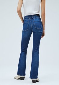 Ausgestellte blaue Denim-Jeans mit hoher Taille, ausgestattet mit zwei Gesäßtaschen und einer glatten Textur. Kombiniert mit hellen Knöchelschuhen.