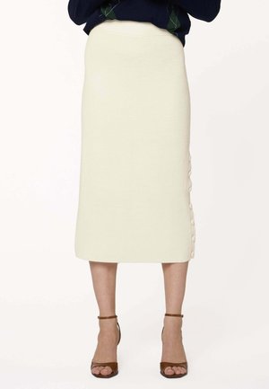 Femme portant une jupe midi côtelée crème avec des boutons latéraux, associée à des talons à bride cheville marron et un haut foncé.