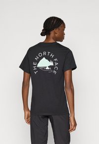 Μαύρο βαμβακερό t-shirt με γραφικό σχέδιο βουνών και δέντρων σε ανοιχτό μπλε, με το κείμενο «THE NORTH FACE» να κυρτώνει γύρω από το σχέδιο στην πλάτη.