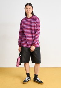 T-shirt à manches longues avec des rayures horizontales rose et violet, associé à un short en denim noir. Baskets noires avec des accents blancs et un sac rose.