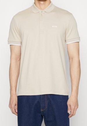 Mann trägt ein beiges kurzärmeliges Polohemd mit "BOSS"-Logo auf der Brust und dunkelblaue Jeans, steht vor einem einfarbigen Hintergrund.
