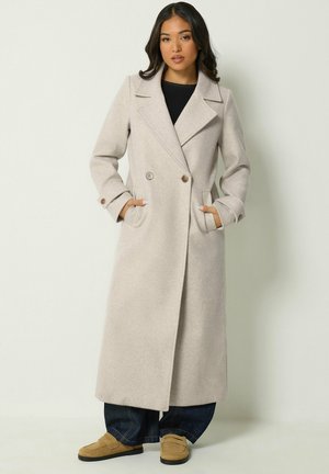 Femme portant un long manteau beige clair à double boutonnage avec de larges revers, un pantalon large foncé et des mocassins marron clair, debout les mains dans les poches.