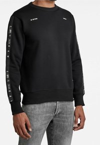 Sudadera negra de mezcla de algodón con cuello redondo acanalado. Presenta un diseño de marca en texto blanco en las mangas y el pecho. Diseño ajustado, mangas largas.