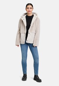Veste à capuche beige avec une texture duveteuse sur le dessus et un motif matelassé en dessous. Présente des boutons-pression et des poches latérales. Portée par-dessus un haut noir.