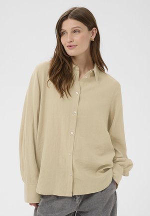 Frau mit langen braunen Haaren, die ein beige-farbenes Hemd mit Knöpfen und graue Jeans trägt, schaut leicht seitlich, die Hände in den Taschen.