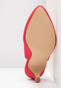Escarpin en suède rouge à talon haut avec un bout pointu et une semelle beige lisse présentant des stries. Positionné sur un fond neutre.