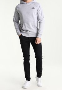 Sudadera con capucha de color gris claro con bolsillo frontal, logo negro en el pecho, combinada con jeans negros ajustados y zapatillas blancas.