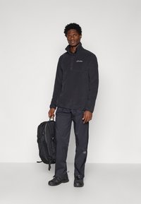 Pullover in fleece nero con zip a un quarto, abbinato a pantaloni impermeabili scuri. Il modello tiene uno zaino nero con spallacci regolabili.