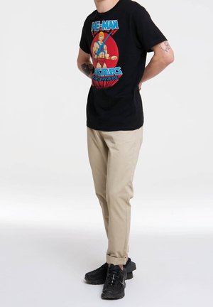 Maglietta nera in cotone con una grafica colorata di He-Man sul davanti, abbinata a pantaloni beige su misura e sneakers nere.