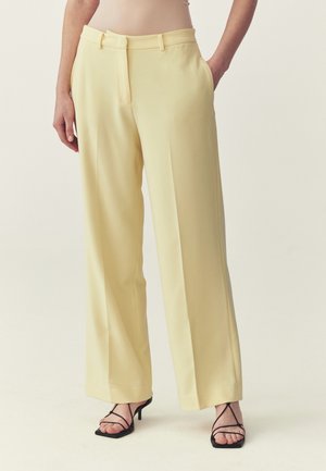 Donna che indossa pantaloni giallo pallido a vita alta e gamba larga abbinati a sandali neri con tacco e cinturini, mani nelle tasche, sfondo neutro.