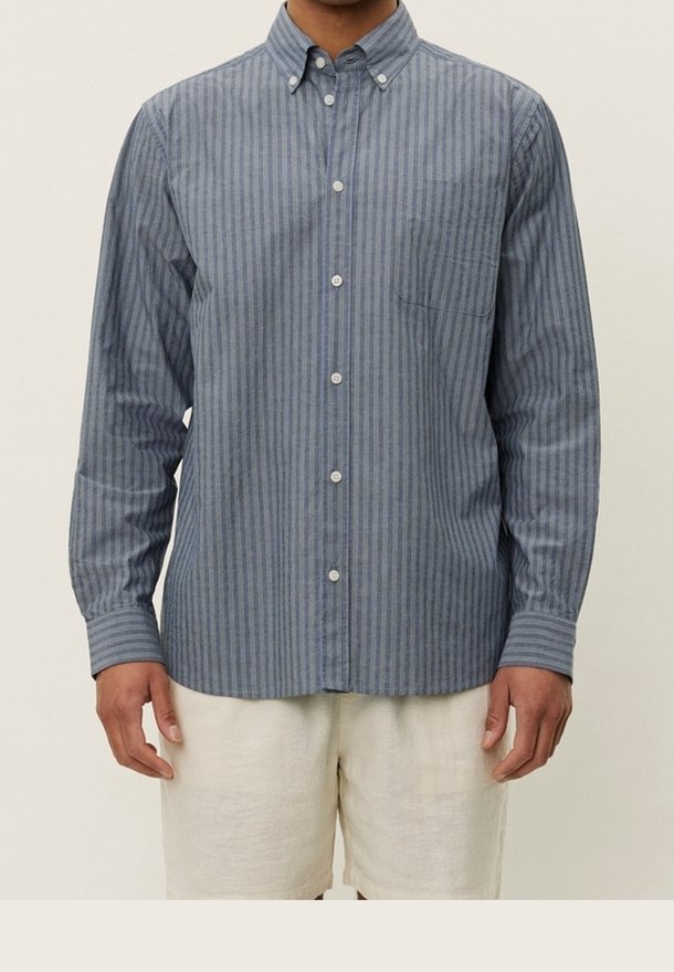 Homme portant une chemise à manches longues bleu et gris à rayures verticales avec un short beige sur un fond uni.