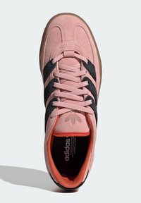 Pink suede sneaker with black three-stripe design, rounded toe, and black rubber sole. Features textured laces and an orange accent near the collar.
