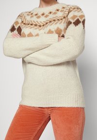 Pull en tricot crème avec un empiècement à motif aux couleurs marron et beige; poignets et ourlet côtelés. Associé à un pantalon en velours côtelé orange.
