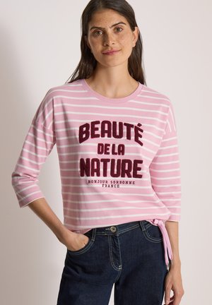 Kvinde med langt mørkt hår iført en lyserød og hvid stribet top med teksten "Beauté de la Nature" og mørke jeans med den ene hånd i lommen.