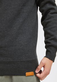 Suéter gris oscuro con dobladillo y puños acanalados. Presenta una etiqueta de cuero marrón cosida en la parte inferior izquierda, textura suave y corte clásico.