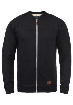 Jūrų laivyno zip-up džemperis, pagamintas iš minkštos medžiagos, su stovinčiu apykakle, dviem priekinėmis kišenėmis ir mažu prekės ženklu odiniu užrašėliu apačioje.