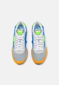 Baskets bleu et gris avec des lacets blancs, des accents jaunes et des logos verts. Le matériau texturé et le col rembourré améliorent le confort.