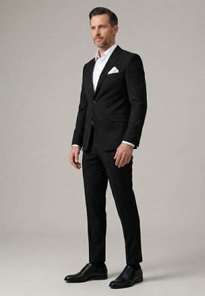 Mann trägt einen schwarzen Anzug, weißes Hemd, weißes Einstecktuch und schwarze Anzugsschuhe, steht vor einem einfarbigen grauen Hintergrund.