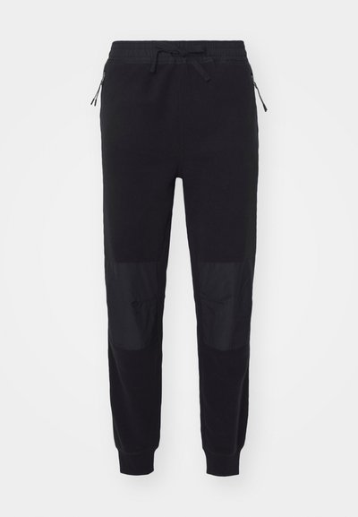 Calças de fato pretas com cintura elástica, cordão, bolsos laterais com zíper e painéis de tecido texturizado nas pernas inferiores.