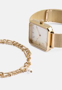 Χρυσό ρολόι με πλέγμα και τετράγωνη πρόσοψη, με μινιμαλιστικό σχέδιο. Δίπλα του υπάρχει ένα χρυσό βραχιόλι αλυσίδας με μια πέρλα.