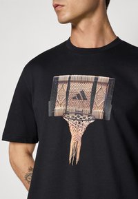Must Must tš-shirt musta värviga, millel on graafika korvpallivõrgu ja kudumisega, millel on nähtav Adidas logo ja keerukad disainielemendid.