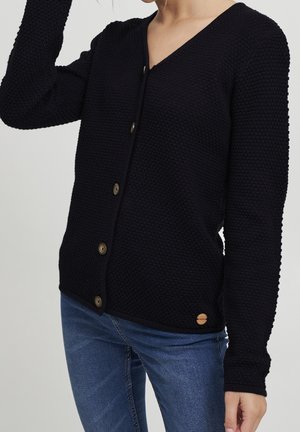 Femme portant un cardigan noir texturé boutonné et un jean bleu, un bras levé vers sa tête.