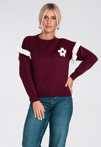 Blondynka w bordowym swetrze z białymi falbanami oraz białą kwiatuszką, w zestawieniu z niebieskimi dżinsami, stojąca z jedną ręką na biodrze.