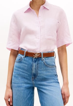 Personne portant une chemise rose clair à manches courtes boutonnée, rentrée dans un jean bleu taille haute avec une ceinture marron et une boucle dorée.