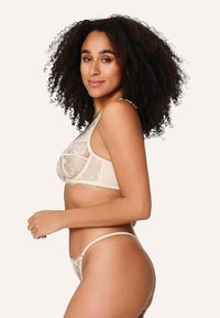 Spitzen-Bralette und -Thong in hellbeige, mit floralen Mustern und transparenten Einsätzen. Zarte Fransenverzierungen entlang der Kanten sorgen für zusätzlichen Textur.