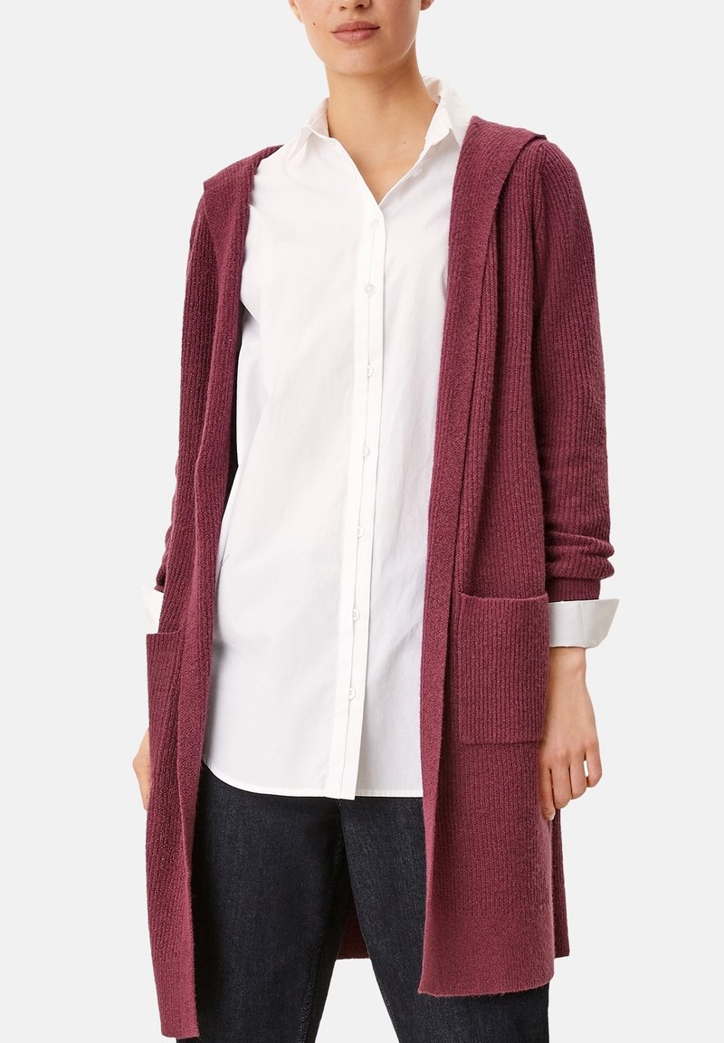 Femme portant un long cardigan tricoté bordeaux ouvert sur une chemise blanche boutonnée et un jean foncé, debout devant un fond blanc uni.
