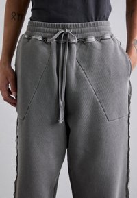 Šedé tepláky s šňůrkou v pase, texturovanou tkaninou, bočními kapsami a geometrickými detaily švů. Doplňky zahrnují kontrastní boční panely.