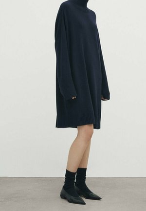 Personne portant une longue robe-pull col roulé oversize bleu marine avec des chaussettes noires et des chaussures pointues noires, debout sur un sol gris.