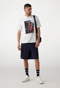 Fehér rövid ujjú póló fekete és piros grafikai mintával, tengerészkék cargo rövidnadrággal, fekete-fehér csíkos zoknival és fehér tornacipővel párosítva.