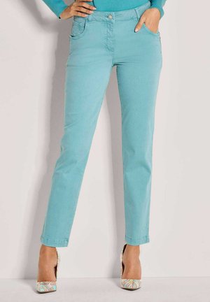 Slim fit light blue pants worn with colorful patterned high heels, hands resting in front pockets against a plain wall.