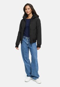 Zwarte hoodie met een ritssluiting aan de voorkant, textuurstof en geribde manchetten, gecombineerd met brede blauwe jeans en witte sneakers.