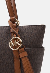 Gros plan d’un sac à main en cuir marron et à motif foncé avec un pendentif logo Michael Kors en or et un pompon en cuir fixé à la anse.