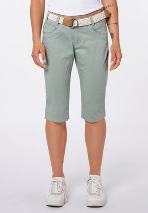 Person trägt kniehohe hellgrüne Shorts, weißen gewebten Gürtel, weiße Sneakers und ein weißes ärmelloses Oberteil und steht mit entspannten Armen.
