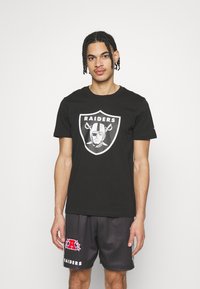 Camiseta de algodón negra con un logo de los Raiders en blanco en la parte frontal. Combinada con pantalones cortos de malla oscuros con letras y acentos en rojo y blanco.