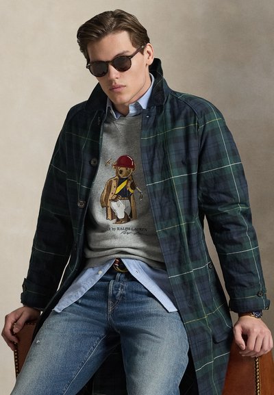 Πράσινο παλτό με καρώ σχέδιο πάνω από γκρι φούτερ με γραφικό αρκούδας, ανοιχτό μπλε πουκάμισο από κάτω και μπλε τζιν. Αξεσουάρ περιλαμβάνουν μαύρα γυαλιά ηλίου.