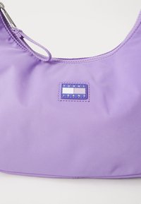 Violetti nailonilaukku, jolla on sileä pinta, kaareva muoto ja ommeltu logotarran, jossa lukee "TOMMY JEANS" edessä.