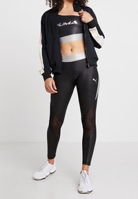 Kvinde iført sort Puma sports-BH, sorte leggings med mesh-paneler, en sort jakke med hvide ærmer og hvide sneakers, stående.