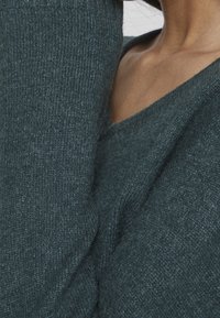 Pull en maille vert sarcelle foncé avec un col en V, doté de poignets côtelés et d'une texture douce. Gros plan détaillé du tissage du tissu.