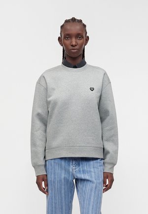 Unge person med flettet hår iført en grå sweatshirt med et lille sort hjerteemblem og blå jeans med striber, stående mod en ensfarvet baggrund.