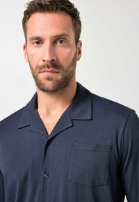 Muž s krátkými hnědými vlasy a vousy, oblečený v tmavě modré košili s límečkem a kapsou na prsou, s neutrálním výrazem, na světlém pozadí.