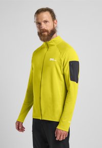 Giacca lime green ad alta visibilità con finitura strutturata, zip completa e tasca con zip nera sulla manica sinistra.