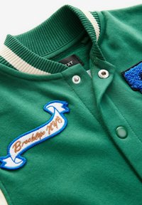 Grön varsityjacka med ribbad krage och krämfärgade ränder. Har en broderad "Brooklyn 19"-patch och tryckknappar. Mjuk tygkänsla.