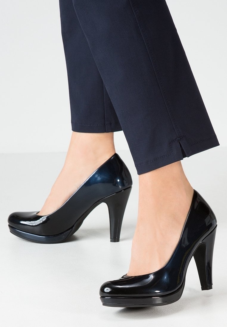 Femme portant des escarpins noirs en cuir verni à talons hauts associés à un pantalon bleu marine longueur cheville avec de petites fentes latérales.