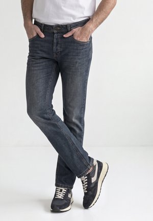 Hombre vestido con vaqueros azul oscuro y zapatillas negras y blancas de pie con las manos en los bolsillos y las piernas cruzadas frente a un fondo blanco.